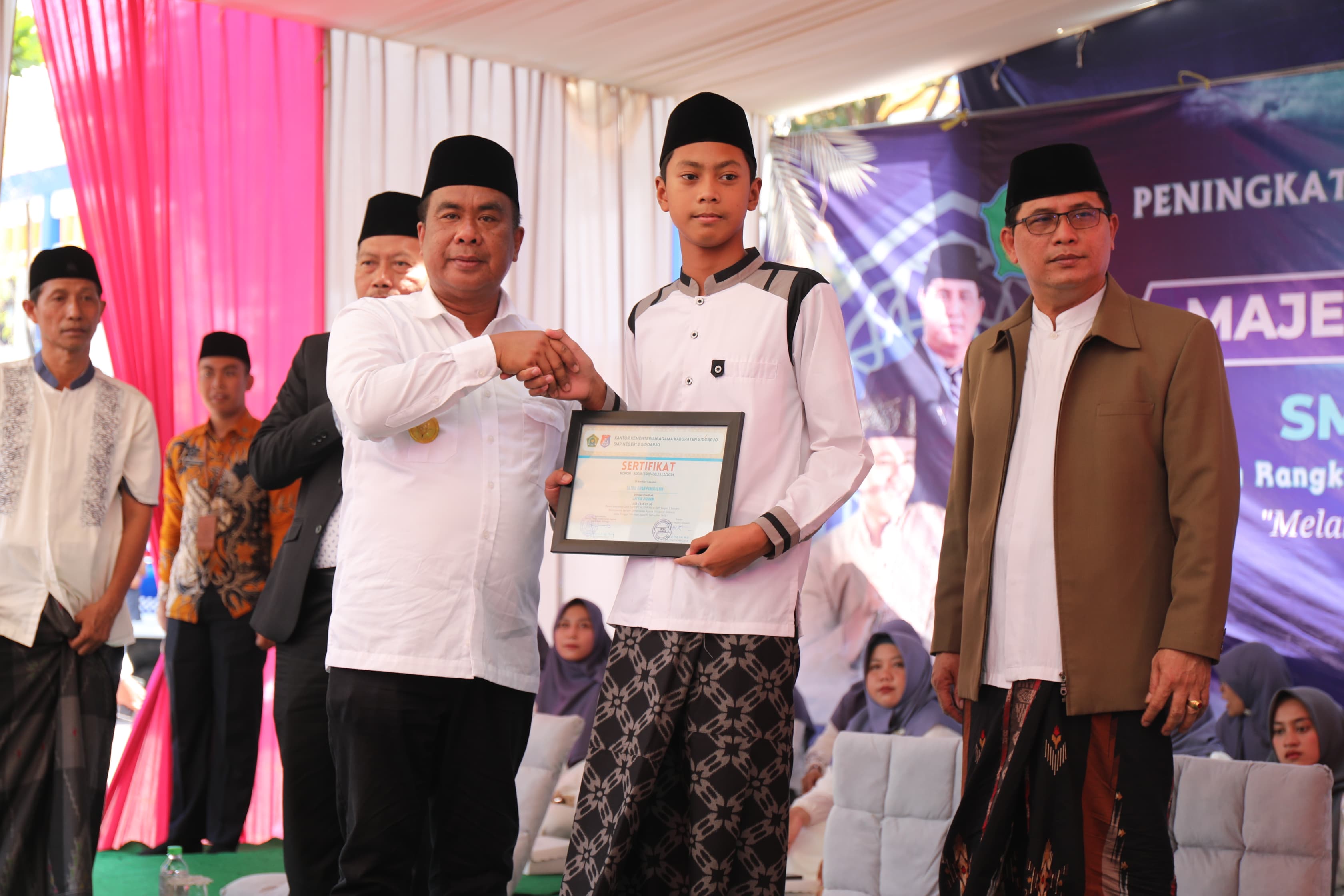 SERAHKAN - Pjs Bupati Sidoarjo, Muhammad Isa Anshori menyerahkan 11 sertifikat hafalan Al Quran kepada para siswa dan siswi SMPN 2 Sidoarjo dalam acara peringatan Maulid Nabi Muhammad SAW yang diselenggarakan di SMPN 2 Sidoarjo, Selasa (01/10/2024).